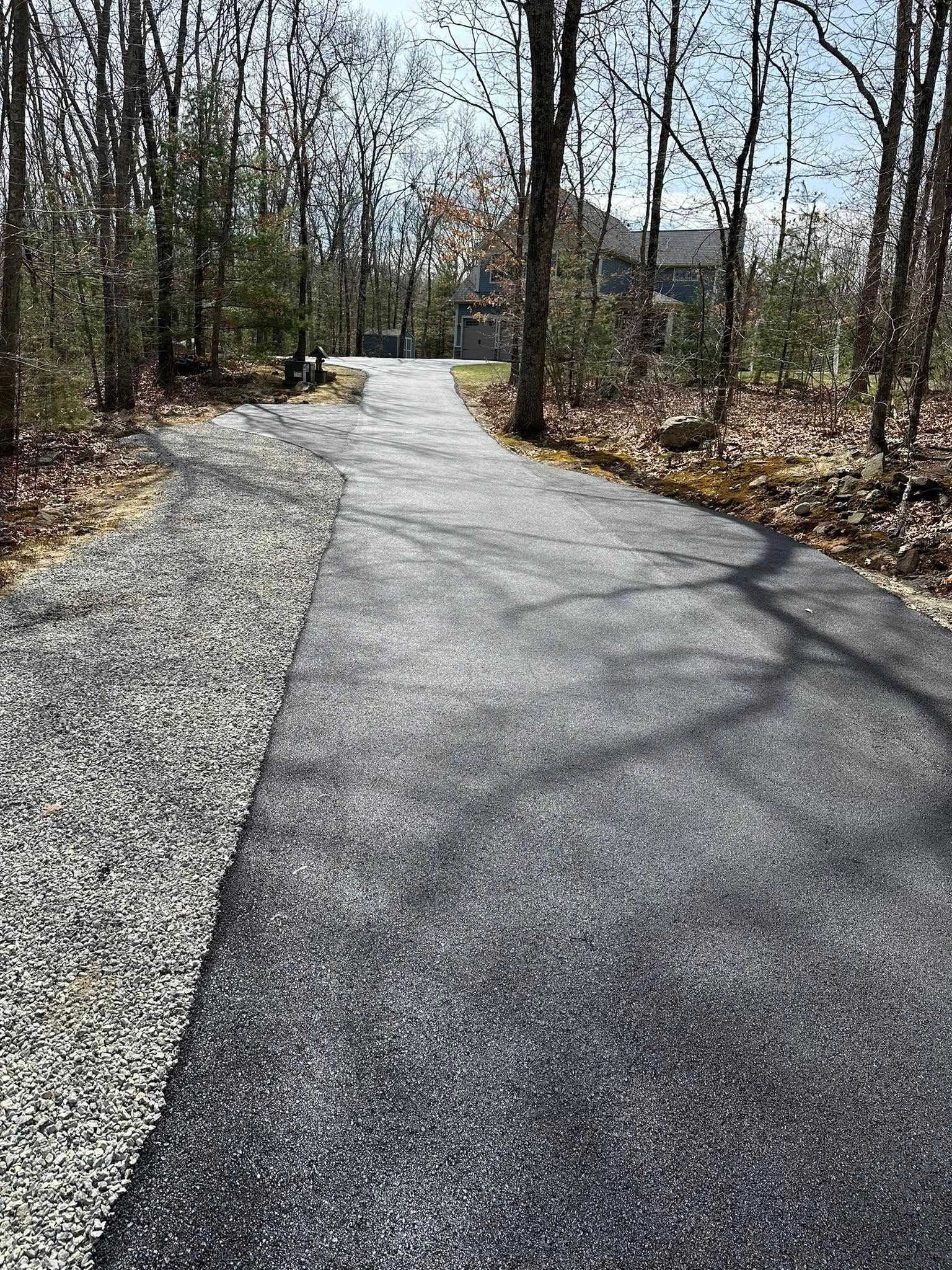 Asphalt driveway with gravel shoulder, trees on either side leading to a distant house.