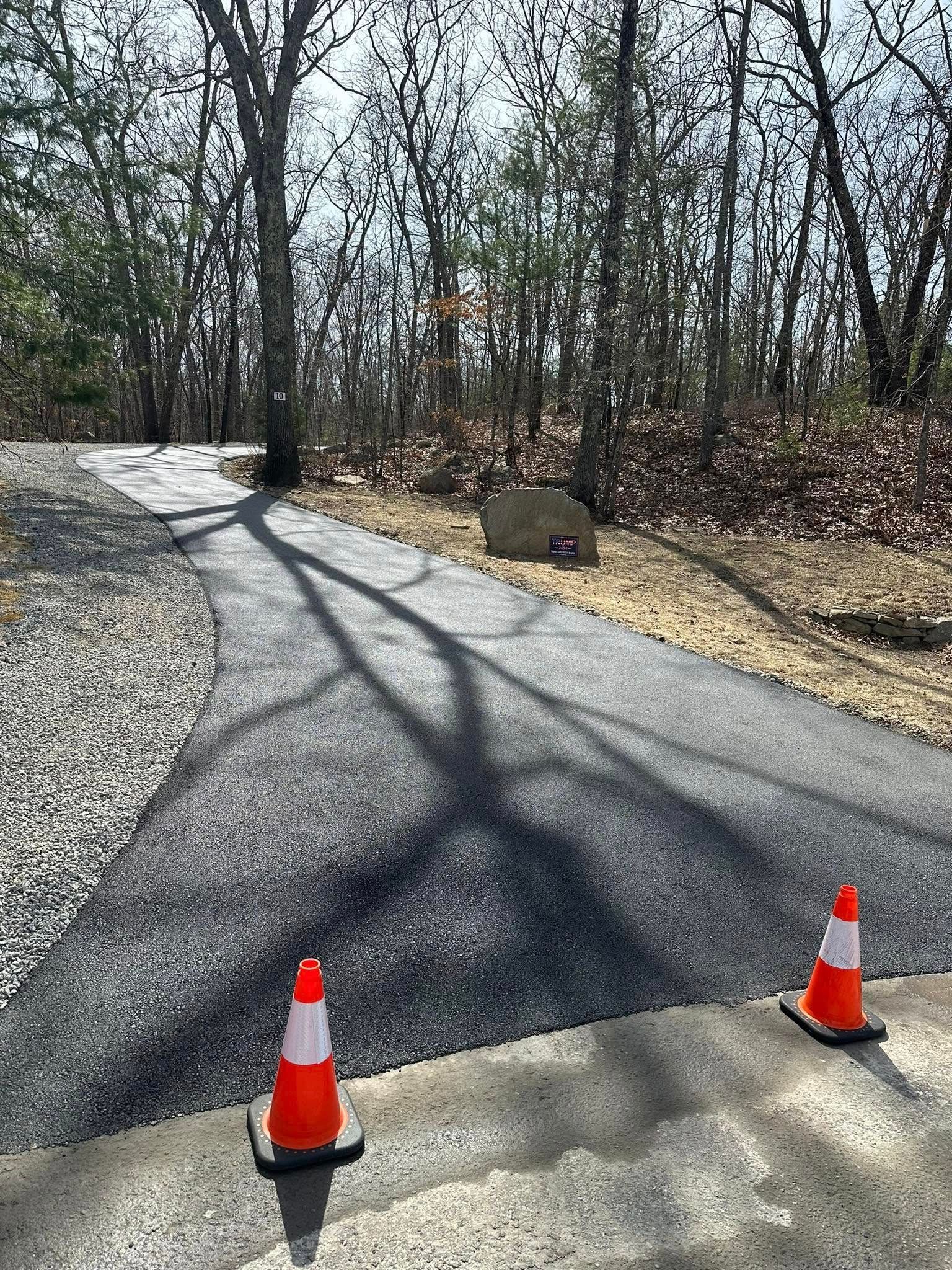 Paved path lined with gravel and orange cones, leading into a wooded area.