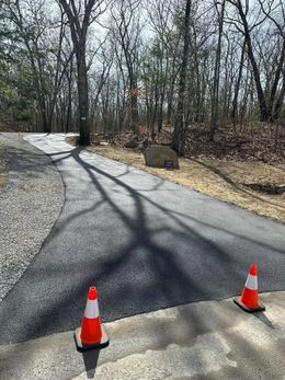 Paved path with orange cones, surrounded by gravel and trees.