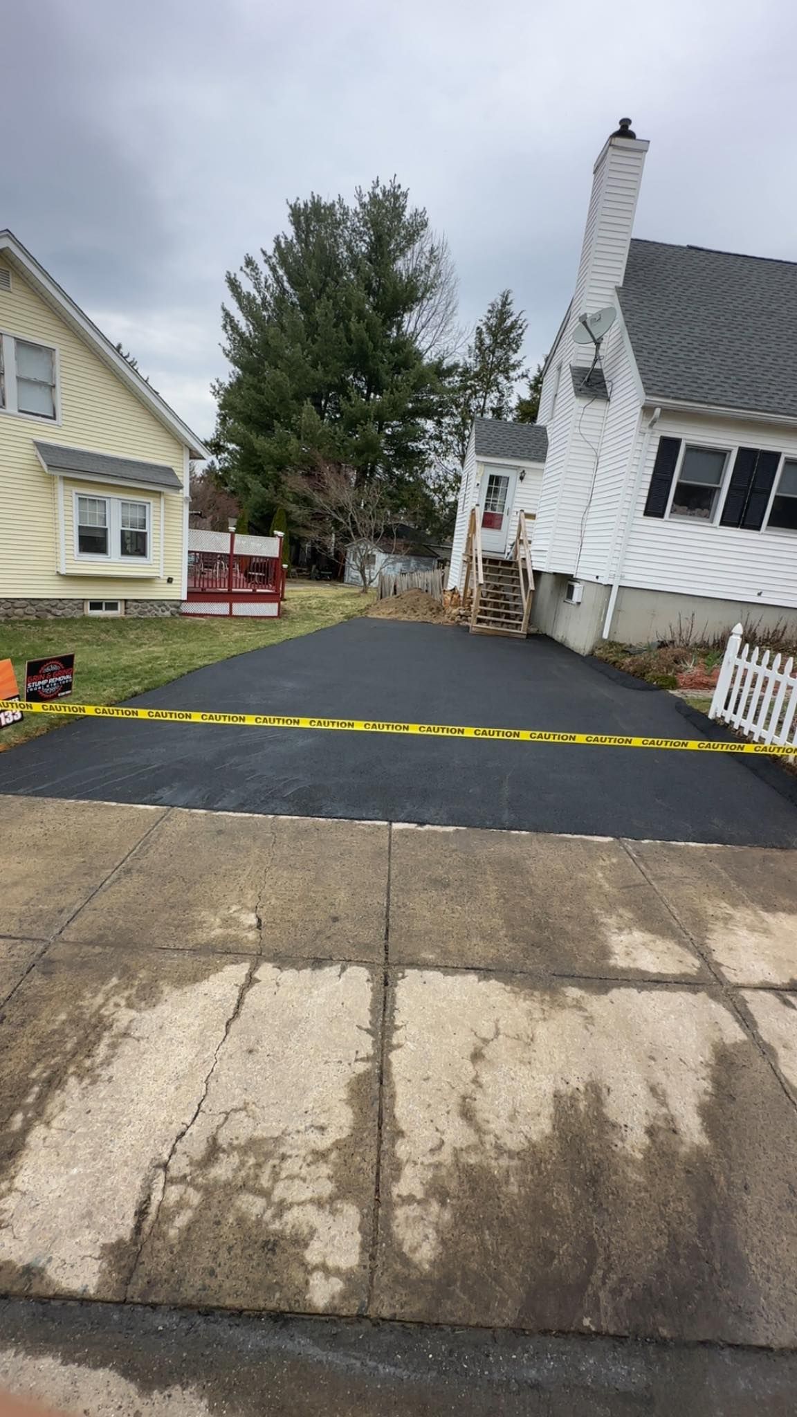 Newly paved asphalt driveway between two houses, blocked by yellow caution tape, overcast day.