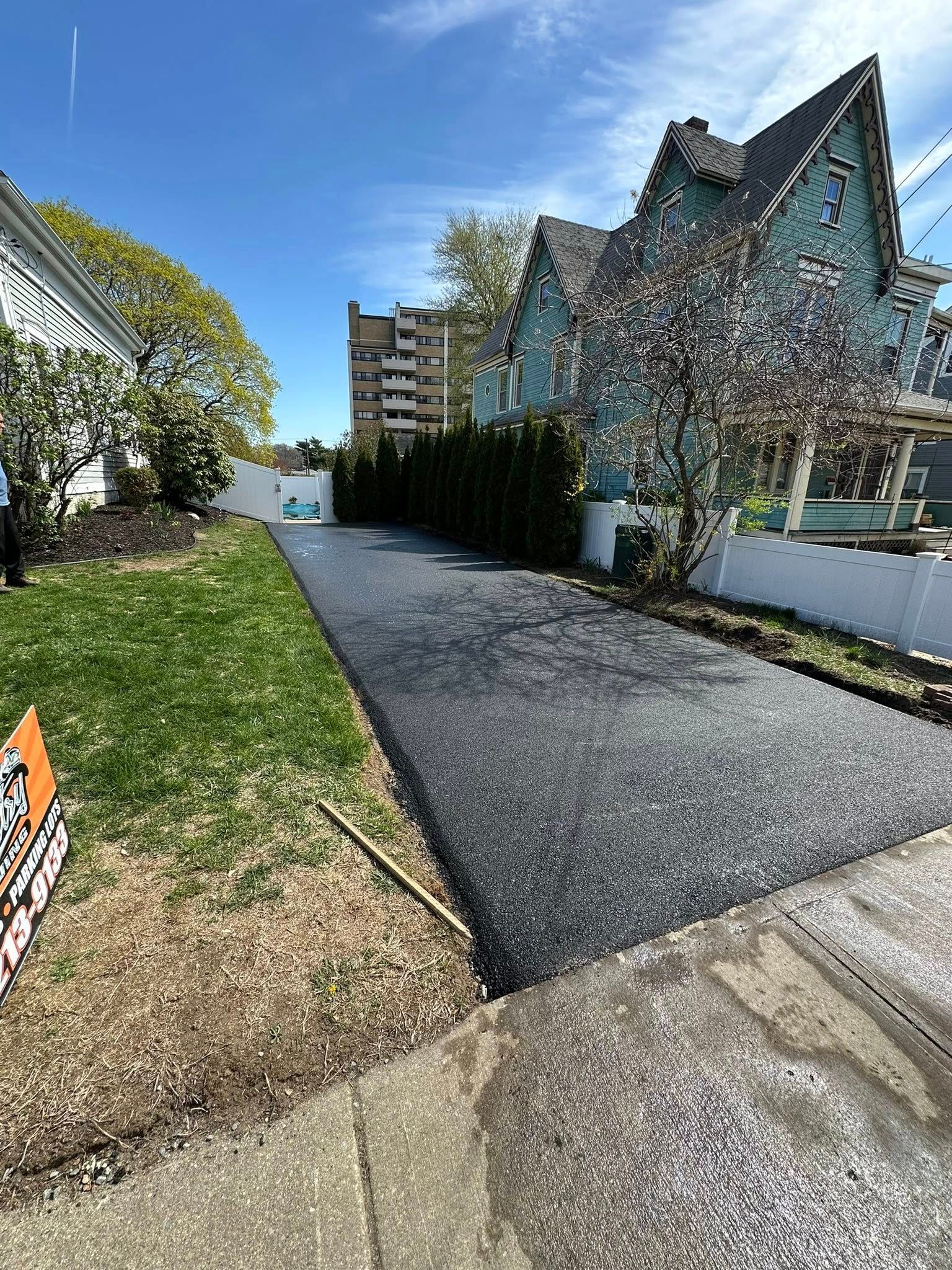Newly paved asphalt driveway between houses, green grass, trees, and blue sky.