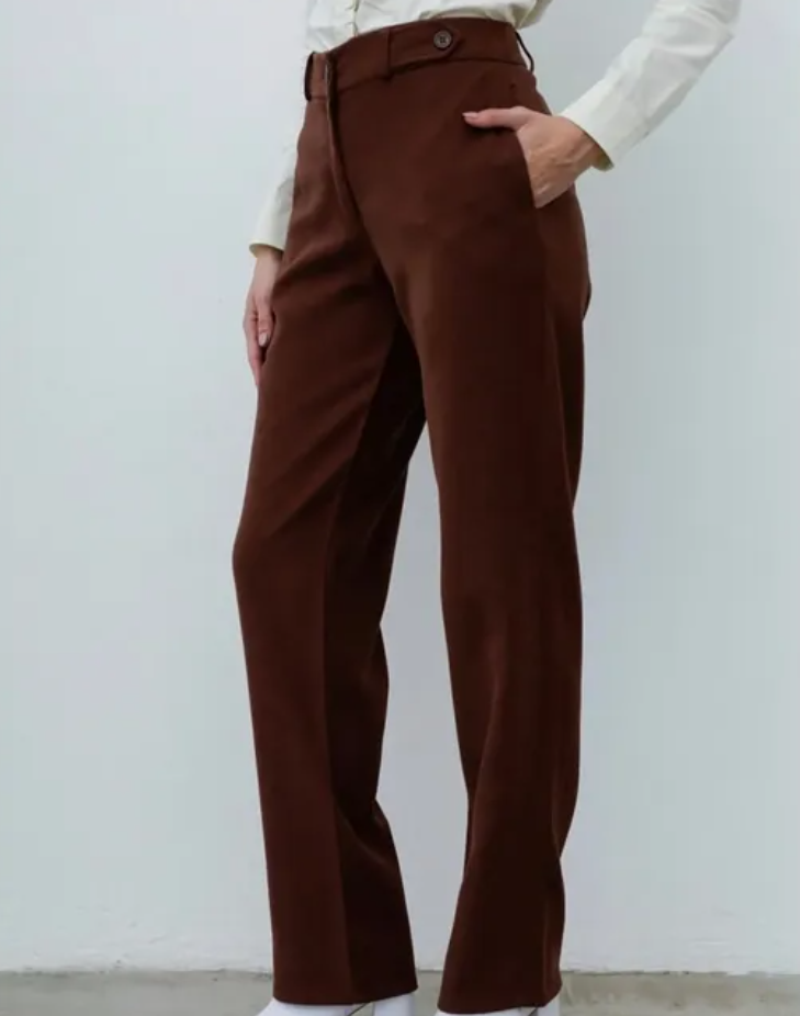 Mujer vestida con pantalones marrones y una camisa de color crema, de pie en el interior.