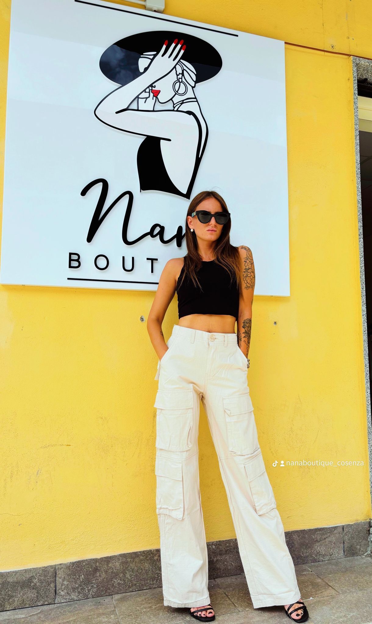 Una donna con un top corto nero e pantaloni bianchi è in piedi davanti a un muro giallo.