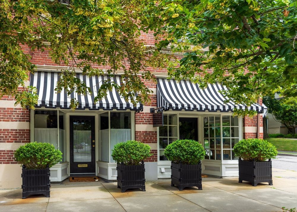 Storefront with black and white striped awnings, red brick exterior, green bushes in black planters.