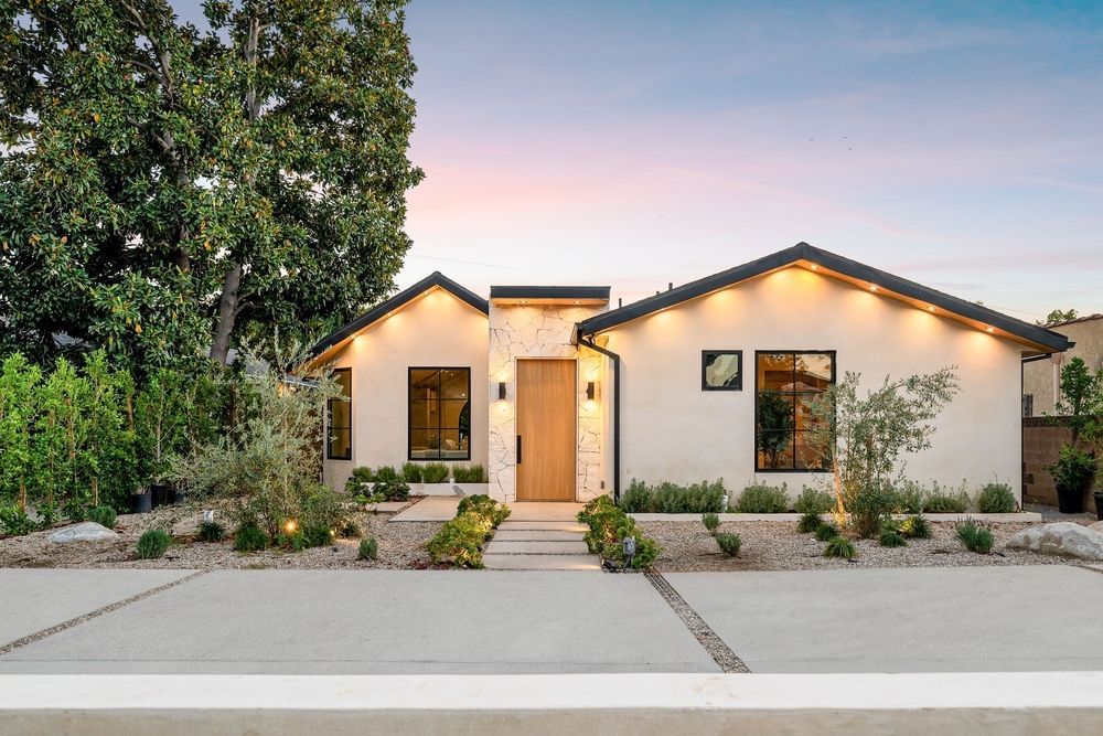 Modern single-story house with light stucco exterior, dark roof, and landscaped front yard at dusk.