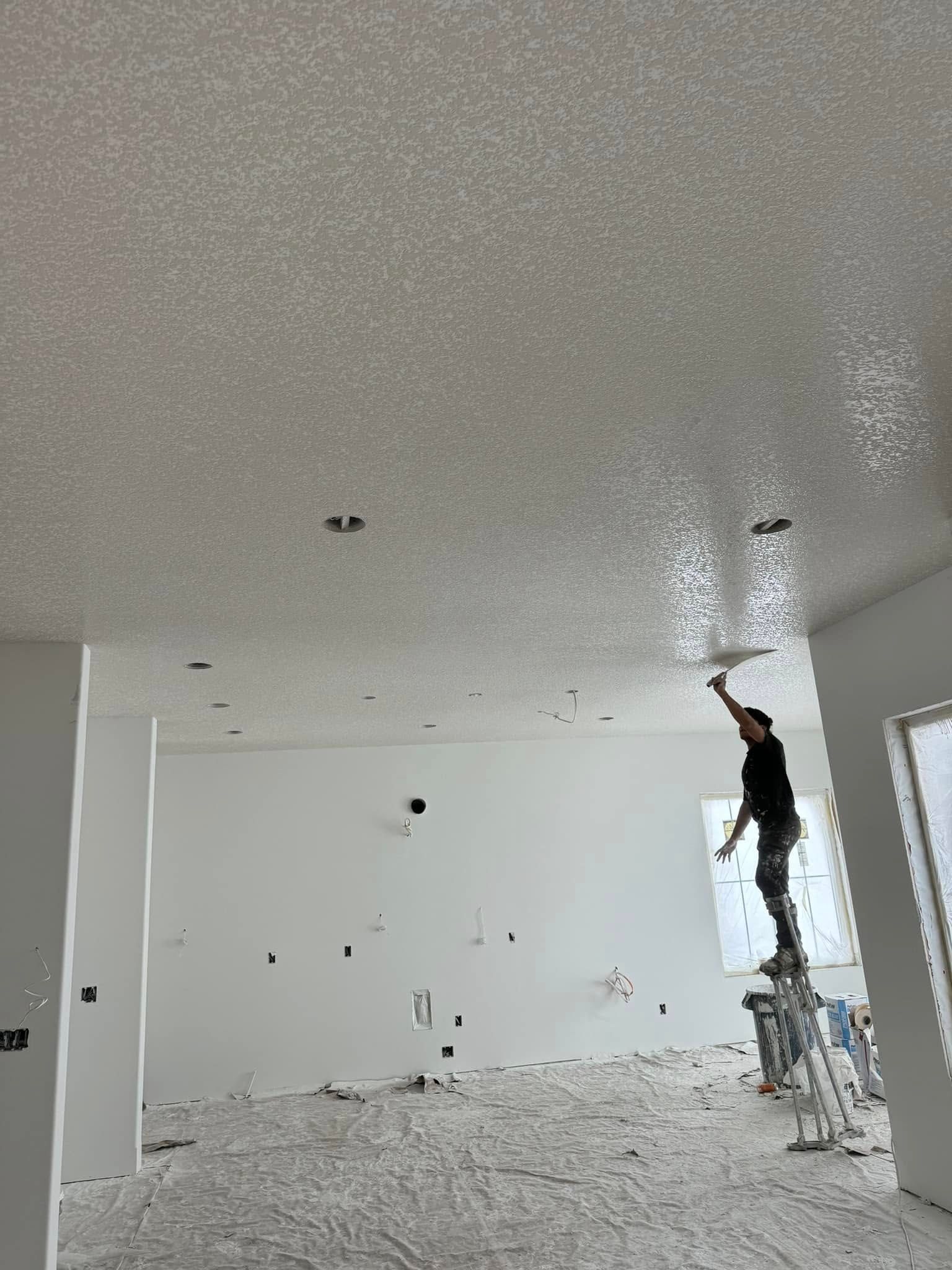 Person on ladder, applying textured ceiling finish in a newly-built room. White walls, debris on floor.