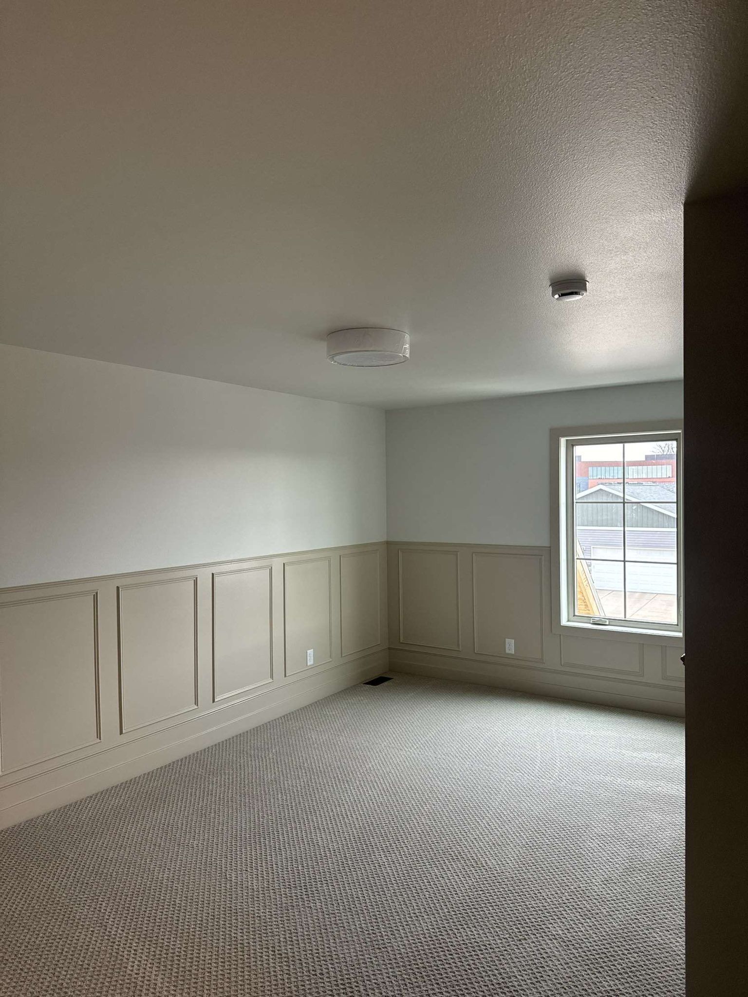 Empty room with white walls, wainscoting, gray carpet, and a window.