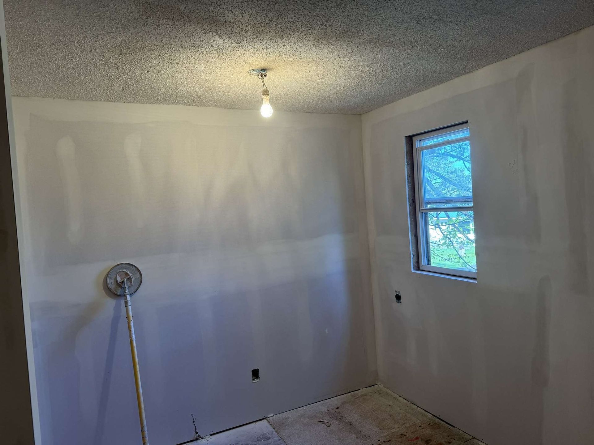 Room with freshly mudded drywall, a small window, and ceiling light. A sanding tool leans against the wall.
