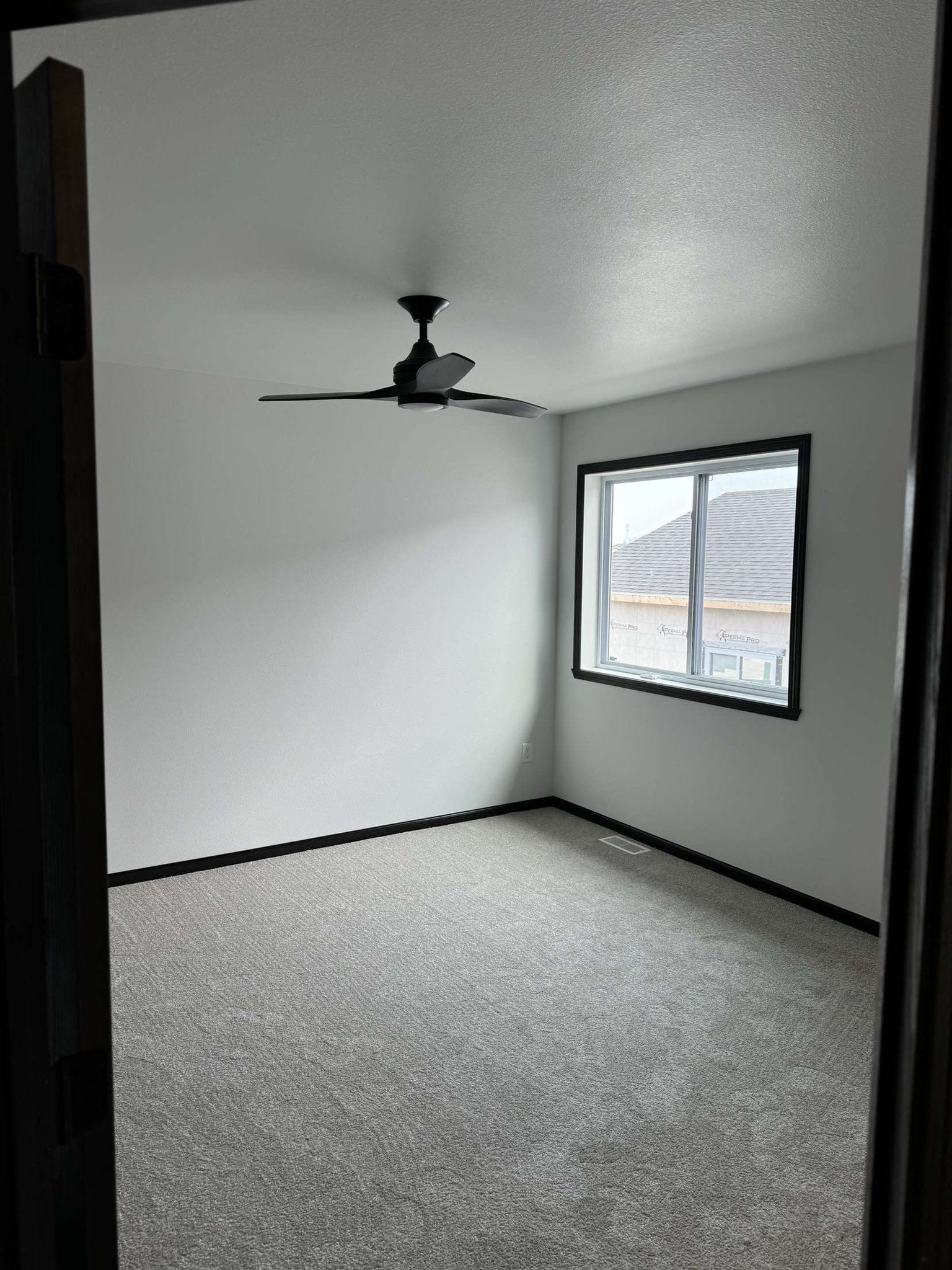 Empty bedroom with gray carpet, white walls, black ceiling fan, and a window.