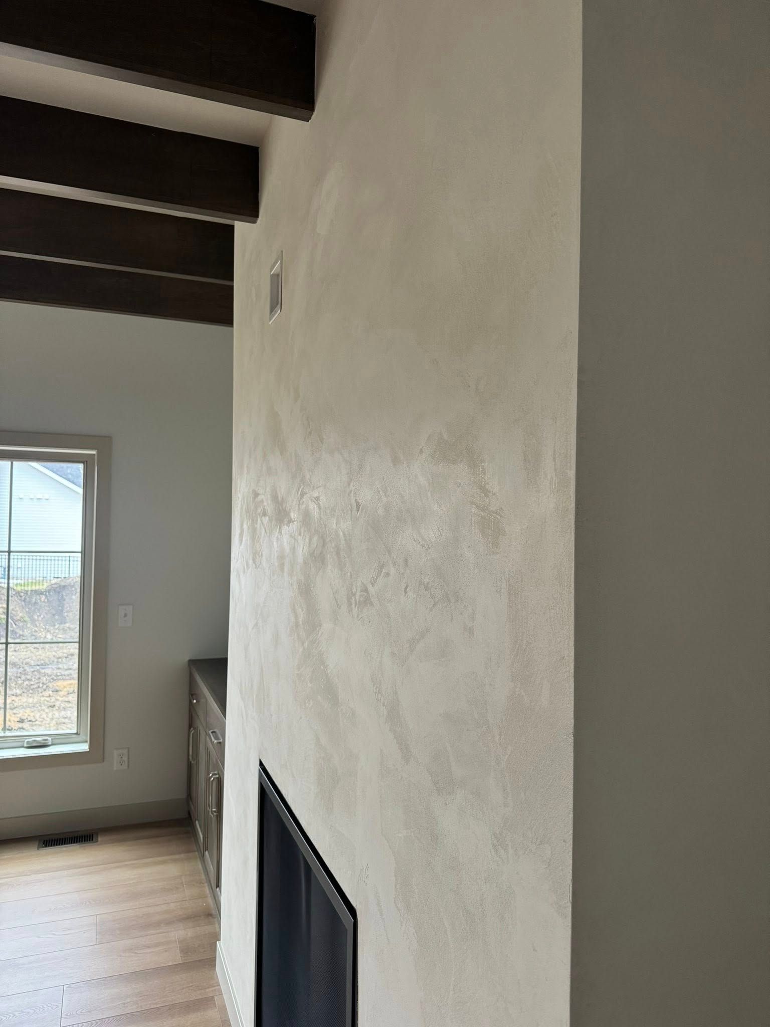 Fireplace with textured, light-colored wall and dark wood beams. Window and wooden floor visible.