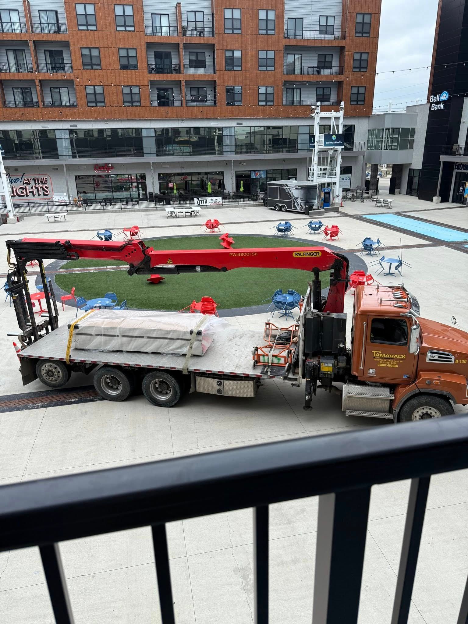 Truck with crane parked in plaza, unloading materials in front of buildings.