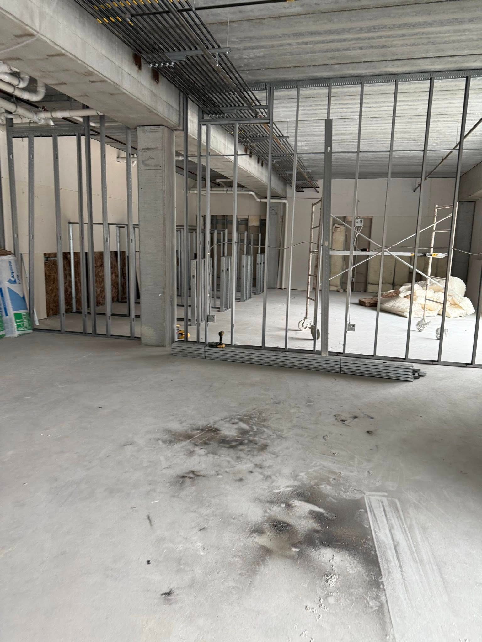 Interior construction site. Metal studs define walls, concrete floor. Overhead wiring visible.