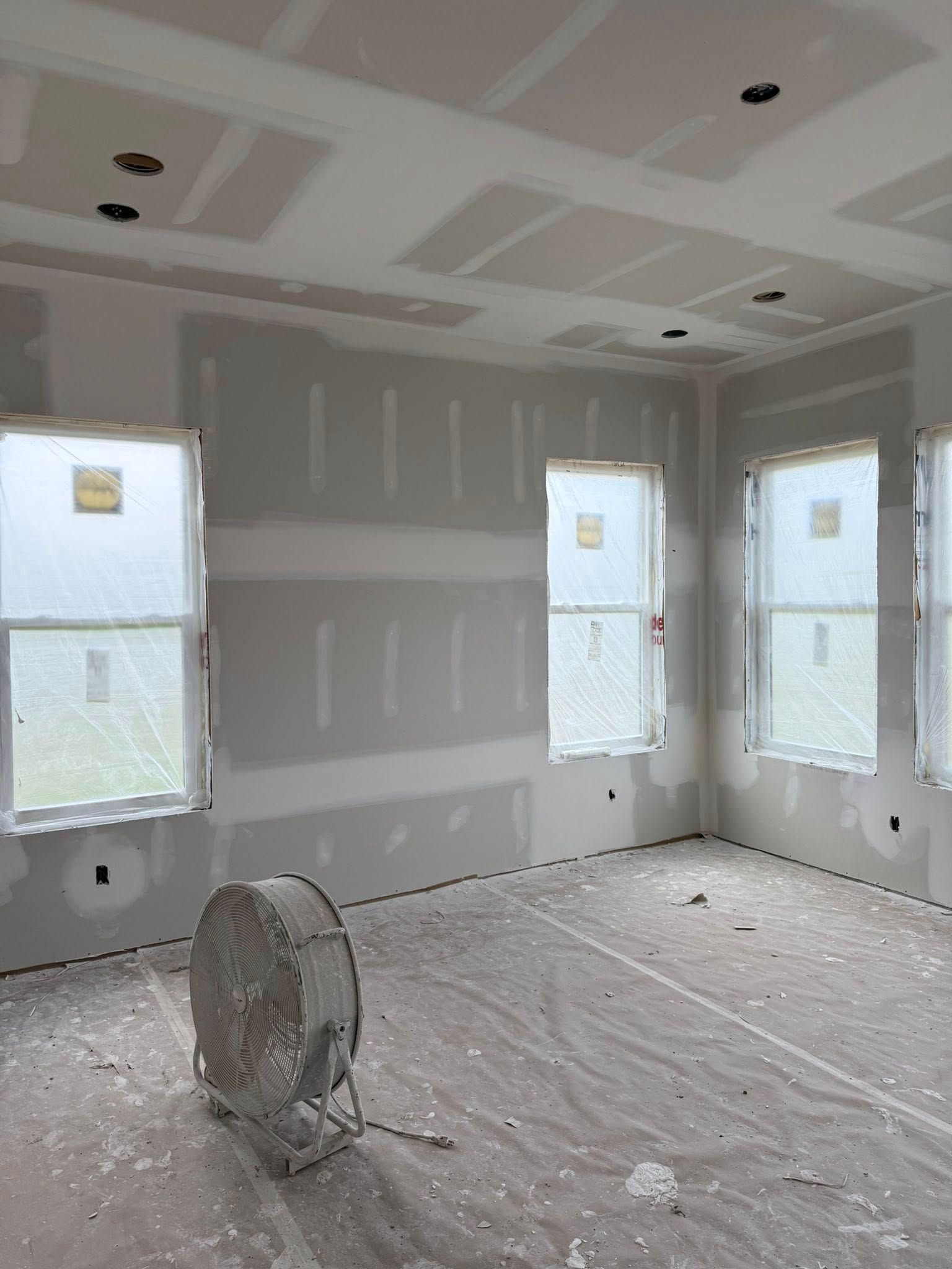 Interior room under construction; drywall, windows, fan, and speckled floor.