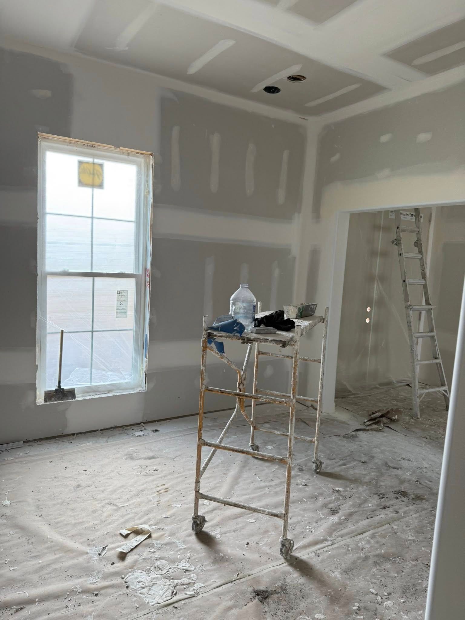 Interior room under construction, drywall installed. Window, scaffolding, ladder, water bottle visible.