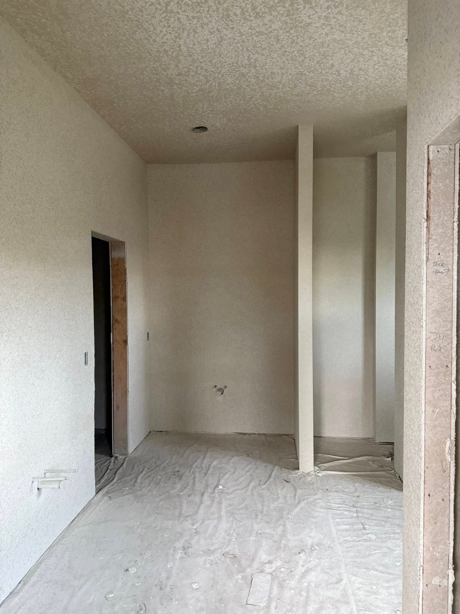 Unfinished interior, likely during construction. Walls and ceiling have stucco texture. An open doorway and vertical wall supports are visible.