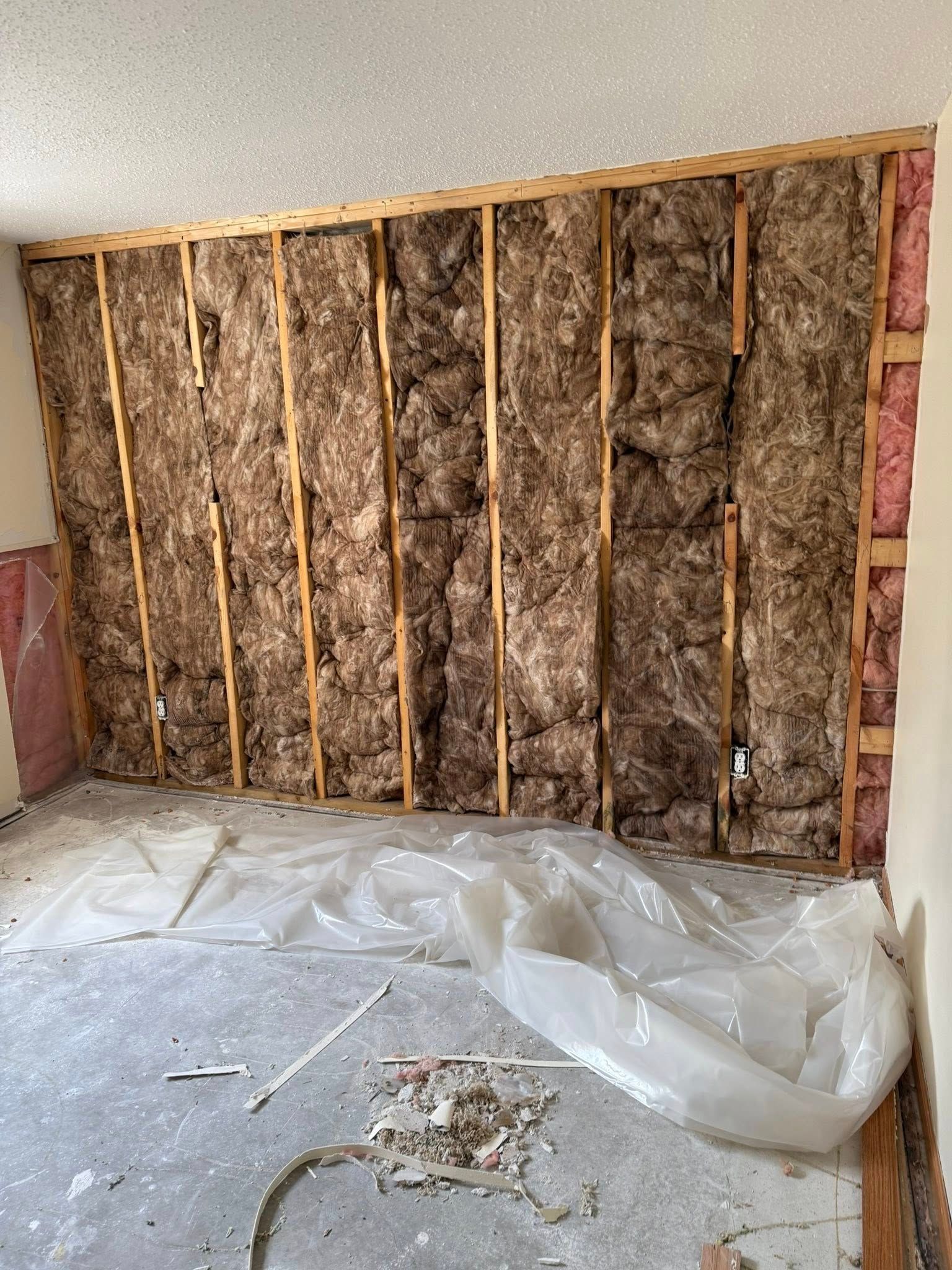 Interior wall with exposed insulation and wooden framing, covered with a plastic sheet on the concrete floor.
