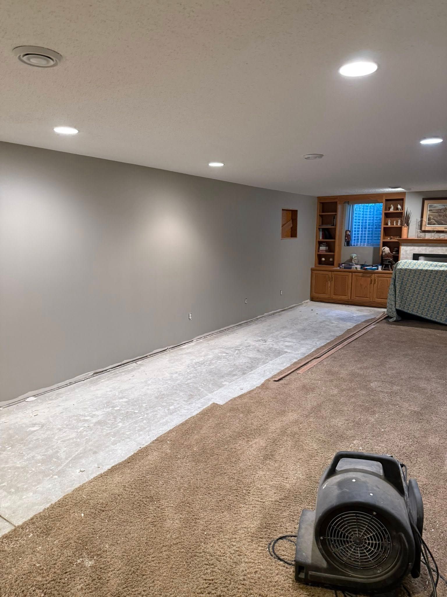 Basement with grey wall, partially exposed floor, brown carpet, and a fan.