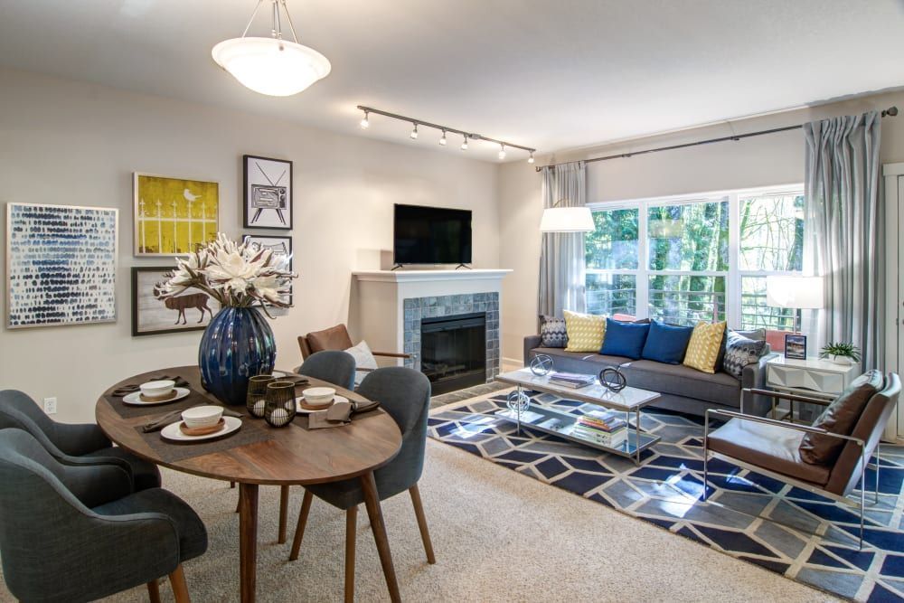 Dining area and living room. Oval table set for two. Gray couch, fireplace, blue and gold rug, large window with view.