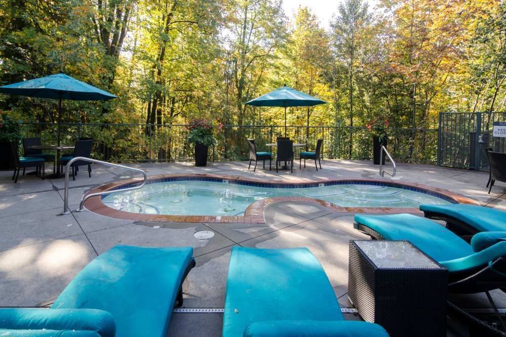 Outdoor hot tub with turquoise lounge chairs and umbrellas, surrounded by trees.
