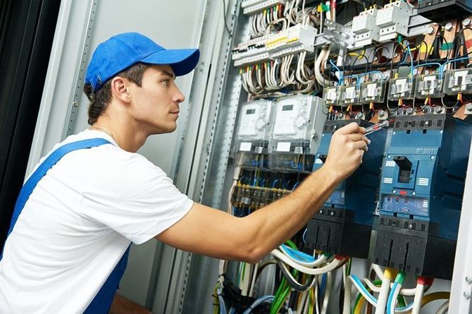 Technician in a blue cap inspects wiring inside an electrical control panel
