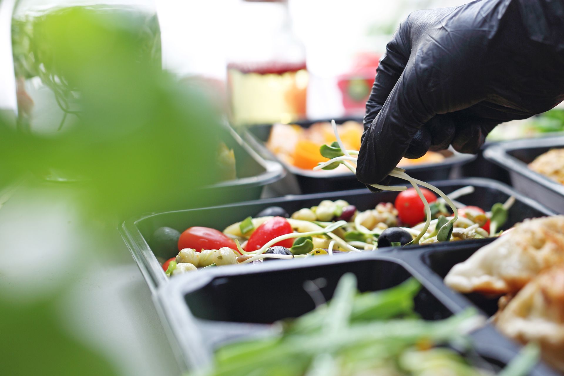 Person in black gloves garnishes a meal prep container with sprouts