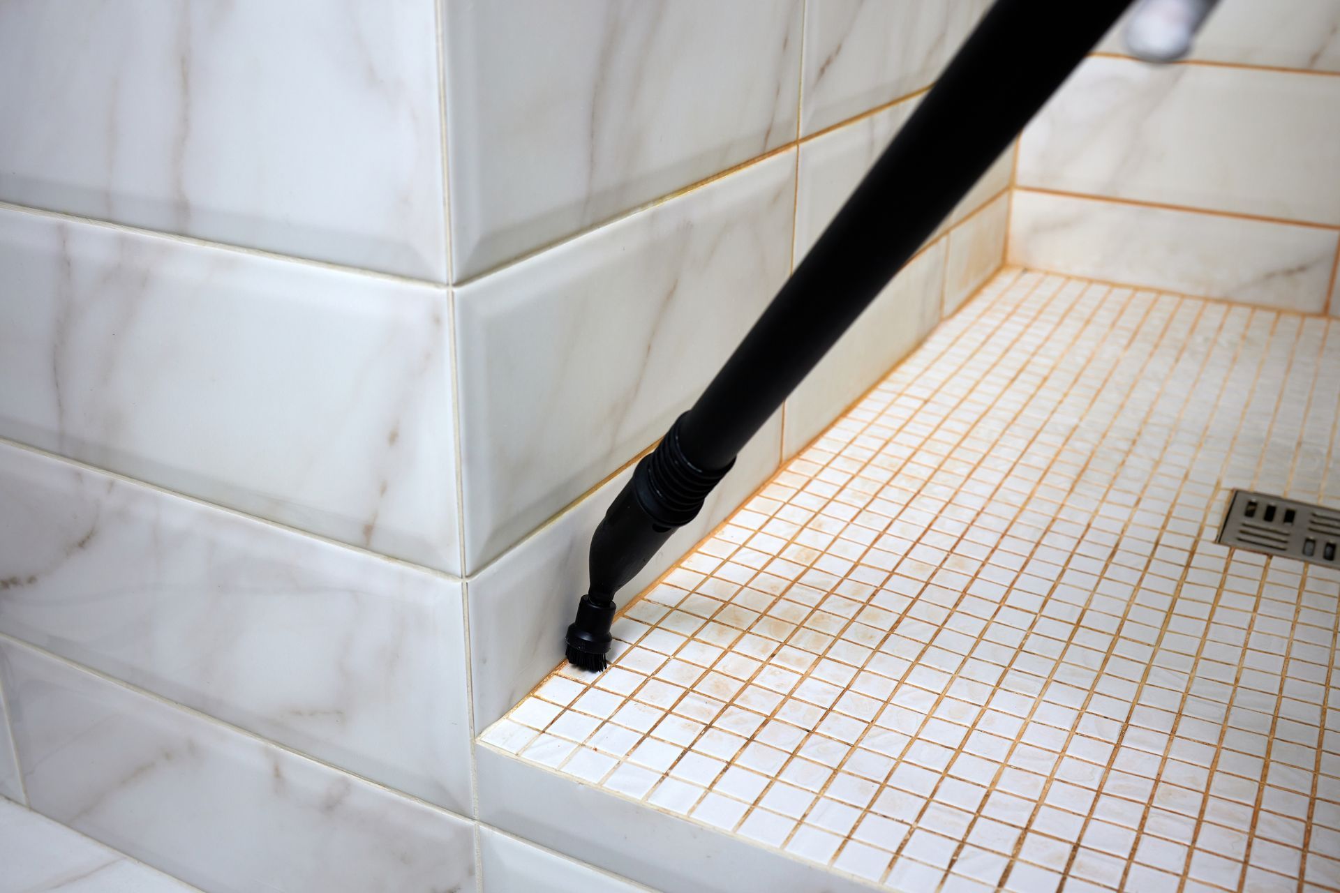 Black steam cleaner nozzle cleaning a white tiled shower floor