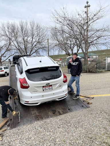 A man is standing next to a white car on a tow truck.
