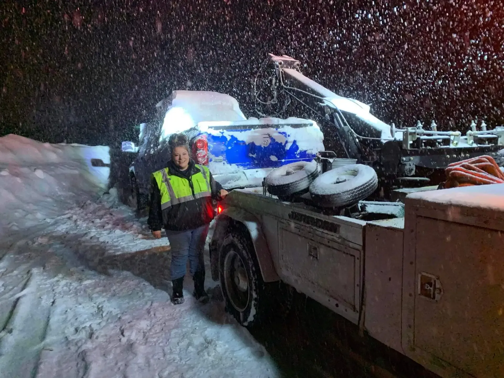 Two people are standing next to a tow truck in the snow.