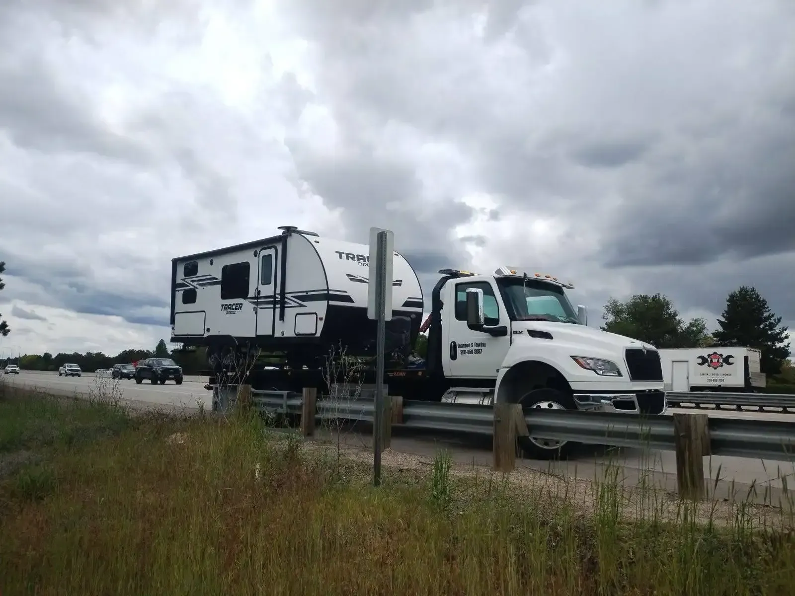 A white tow truck is carrying a trailer down a highway