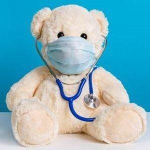 A cream-colored teddy bear wears a blue medical face mask and has a blue stethoscope draped around its neck.