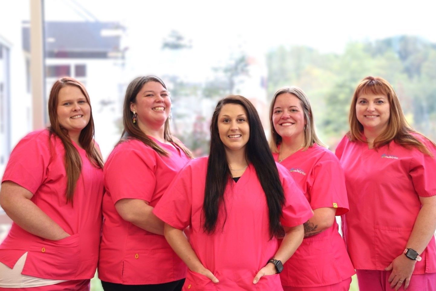Five professionals in bright pink scrubs stand side-by-side smiling in a brightly lit office setting.