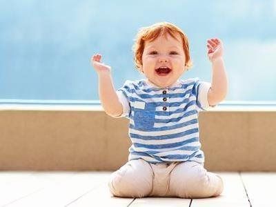 A toddler with ginger hair, wearing a striped shirt, sits on a deck and smiles with their arms raised.