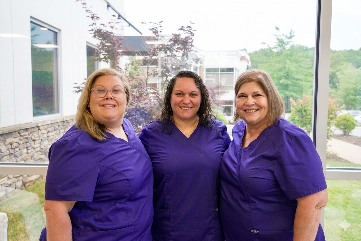 Three professionals in matching navy blue medical scrubs smiling and standing together in a bright, sunlit indoor setting.