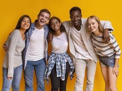Five diverse young people standing together in a line against a solid yellow background, smiling and embracing.