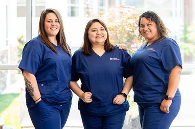 Three professionals in matching navy blue medical scrubs smiling and standing together in a bright, sunlit indoor setting.