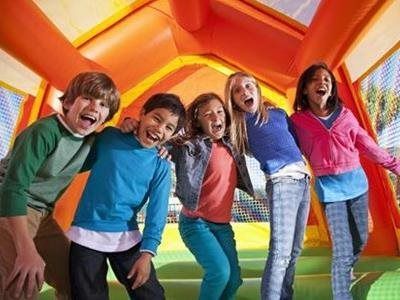 Five children laughing and smiling while standing together inside a bright orange and yellow inflatable bounce house.