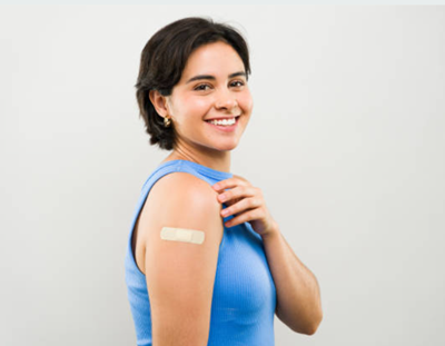 A smiling person in a blue tank top points to a bandage on their upper arm against a plain gray background.