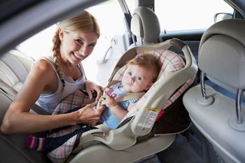 A person smiling while fastening the harness of a baby in a car seat inside a vehicle.