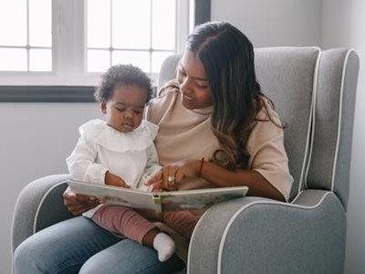 A person sits in a grey armchair, holding a young child on their lap while reading an open book together.