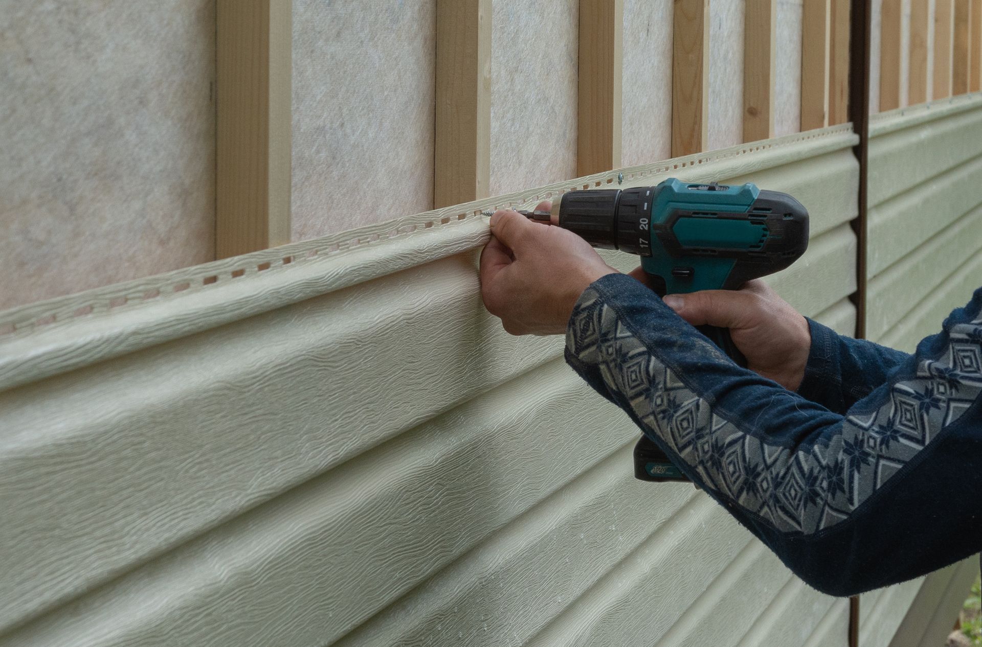 A person installing beige vinyl siding on a building exterior using a cordless drill.