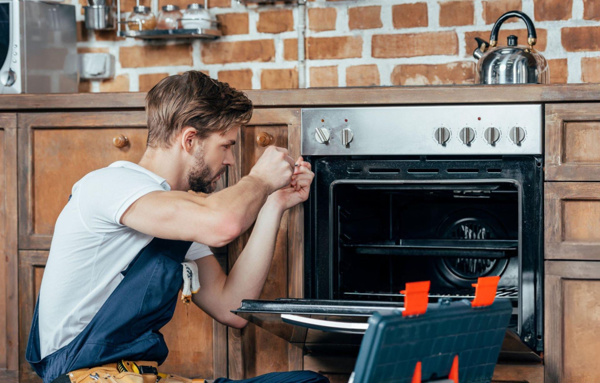 Young technician in protective workwear, performing appliance repair in a residential kitchen.