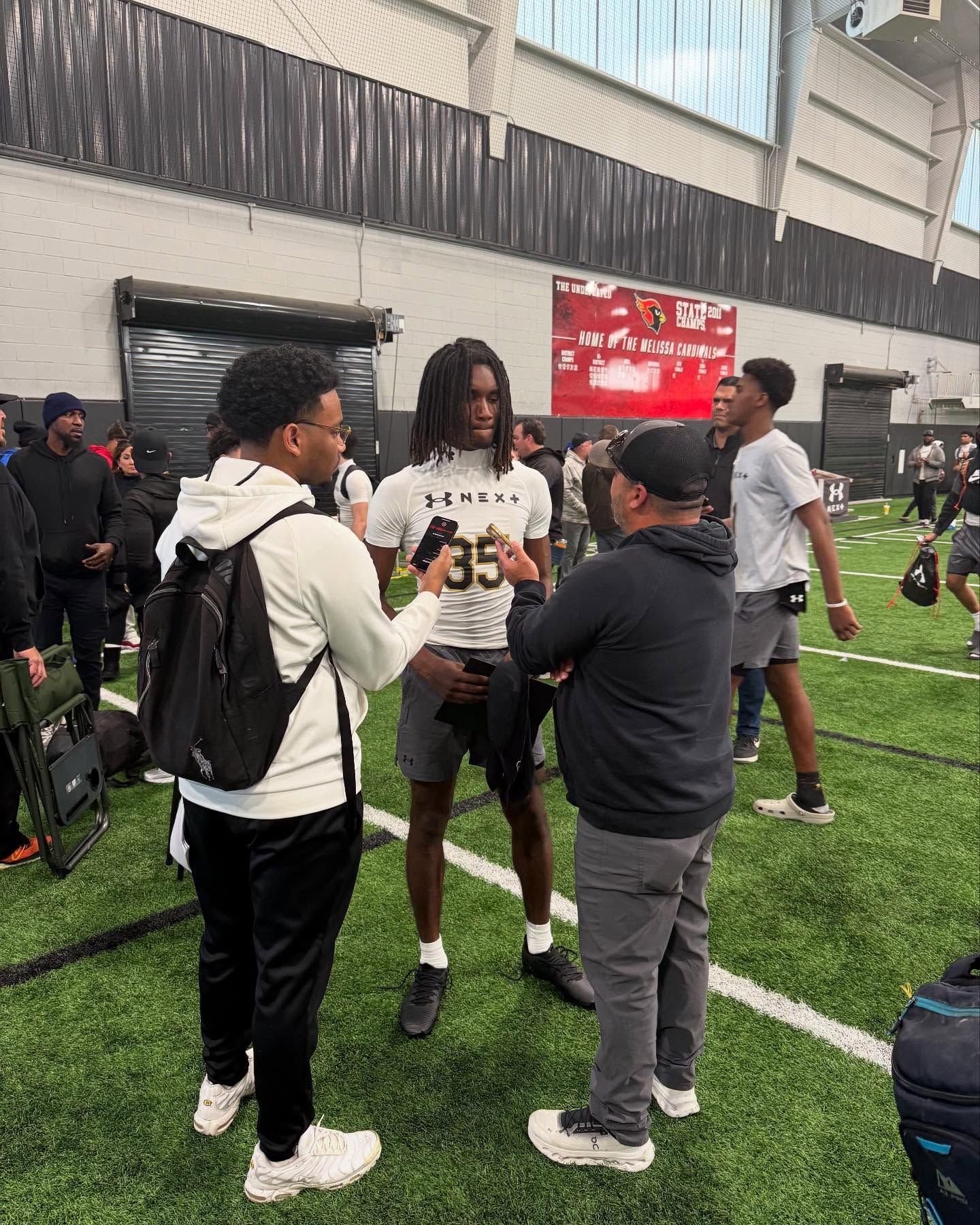 Smiling person with a shaved head, glistening skin, and a gold chain; black background.