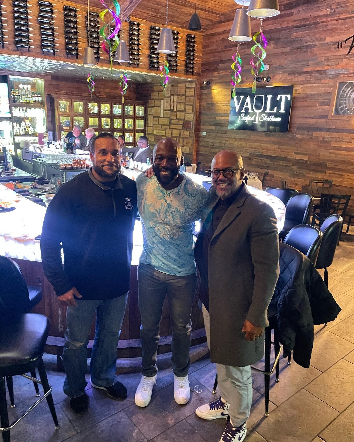 Three men smiling, posing at bar in Vault restaurant. Wooden bar, decor, and sign visible.