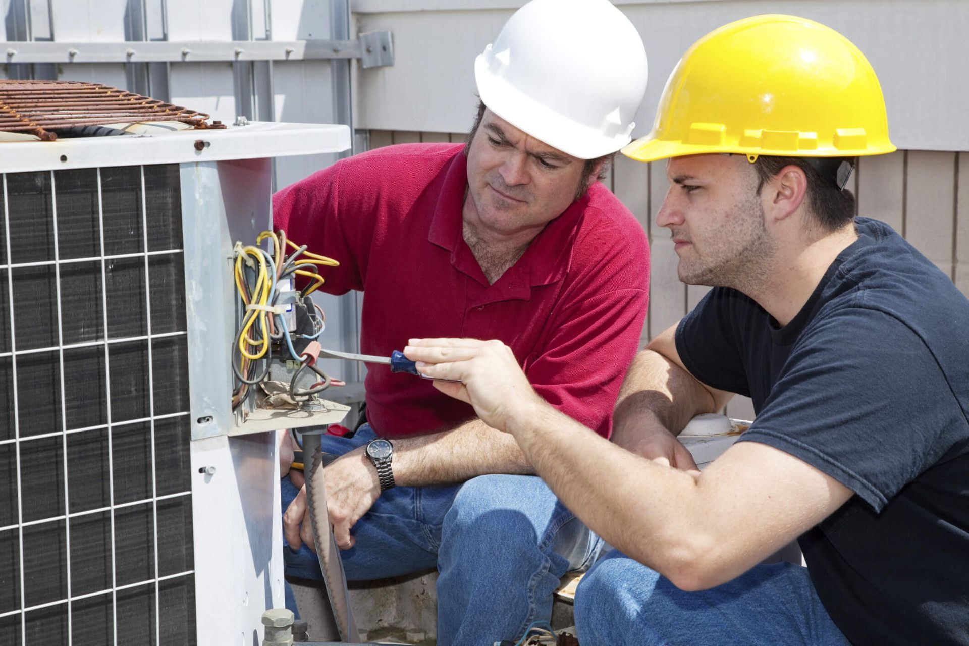 Two men wearing hard hats are working on an air conditioner.