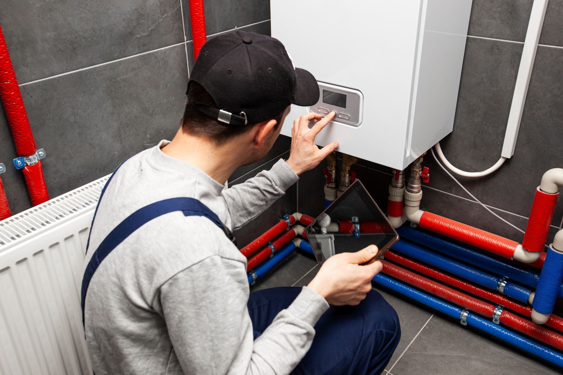 A man is kneeling down in front of a boiler and looking at a tablet.