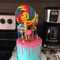 Colorful lollipop-topped cake with pink drips on a kitchen counter near a coffee maker