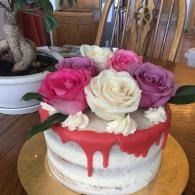 White cake with red drip icing and pink, cream, and purple roses on top on a wooden table