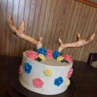 White cake with colorful flowers and antler decorations on a dark table against a wood-paneled wall