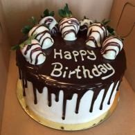 Chocolate birthday cake with white icing drips and strawberries, topped “Happy Birthday” in a bakery box