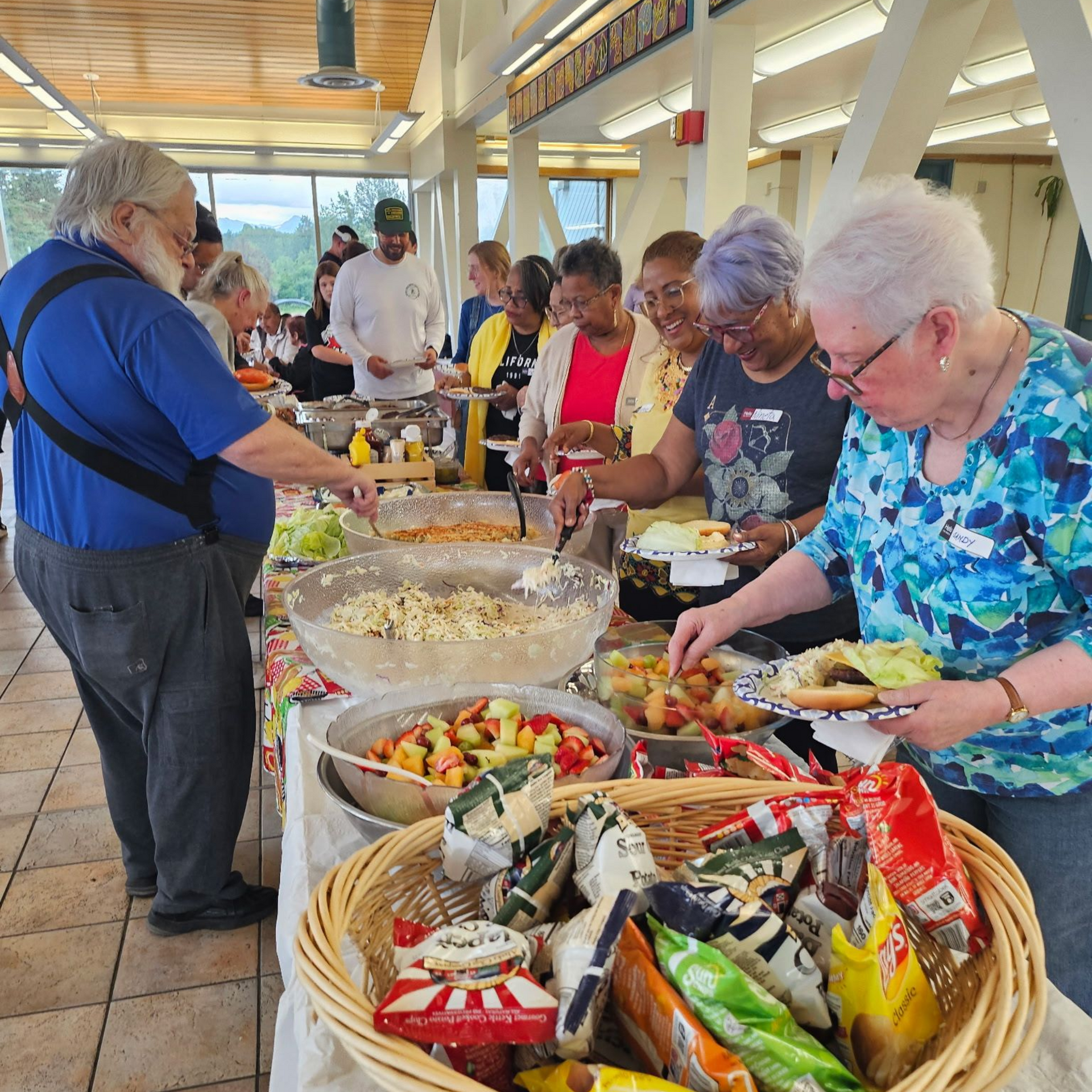 People at a buffet table, selecting food. Bowls of salads, snacks, and condiments. Bright, indoor setting.